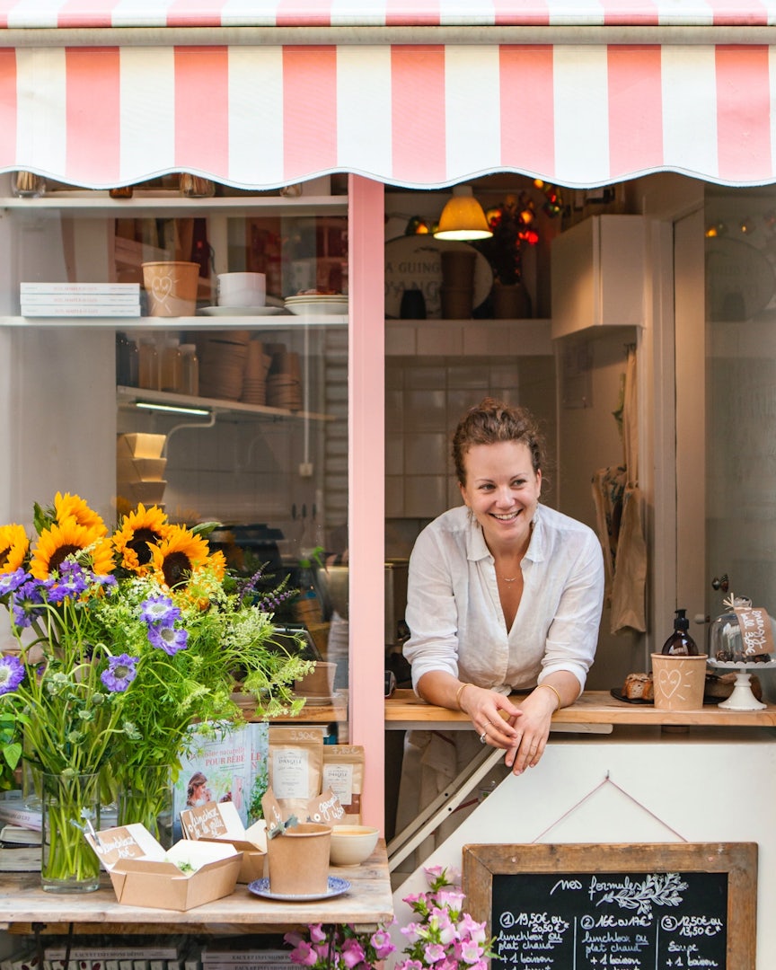 A waitress leans over the counter at La Guinguette d'Angèle in Paris with its white and pink awning and sunflowers on the sidetable.